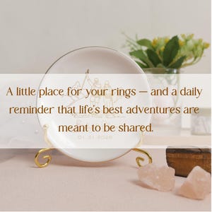 May include: White ceramic ring dish with gold trim and a gold stand. The dish features a gold illustration of two figures and the text: "A little place for your rings - and a daily reminder that life's best adventures are meant to be shared. 01.31.2026."