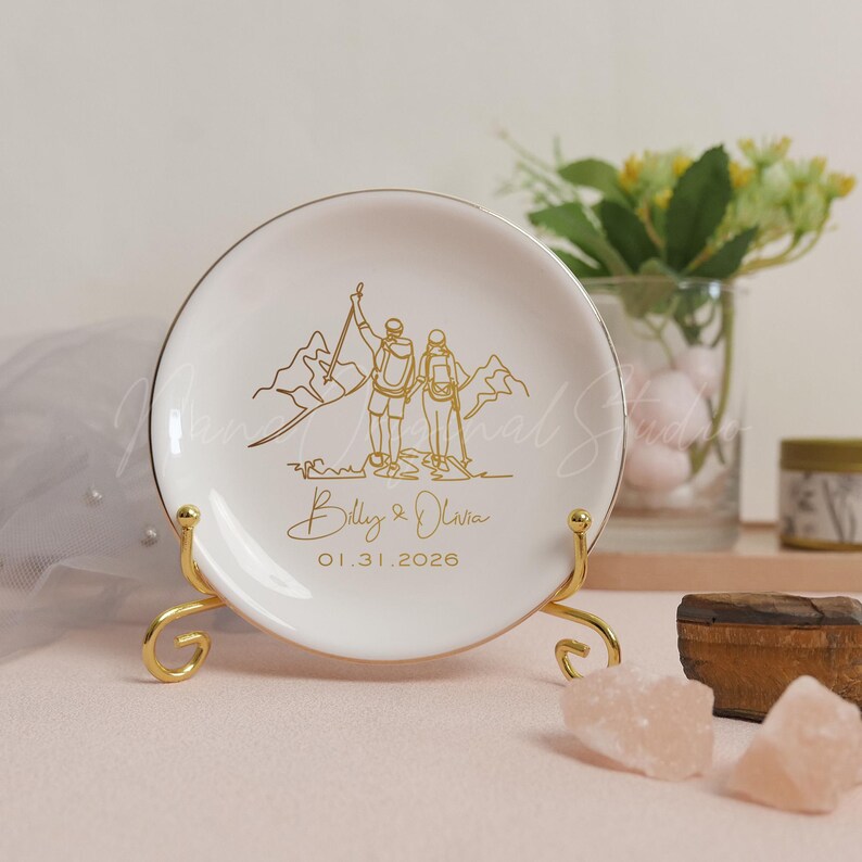 May include: A white ceramic plate with a gold rim, featuring a gold line drawing of two hikers on a mountain. The plate is inscribed with the names "Billy & Olivia" and the date "01.31.2026". The plate is displayed on a gold stand.