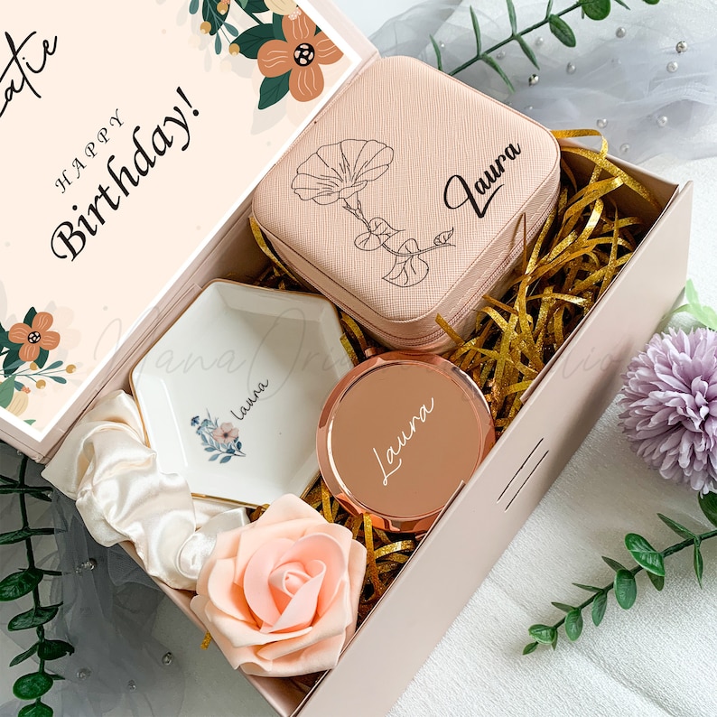Puede incluir: Una caja de regalo rosa con un mensaje personalizado que dice "&iexcl;Feliz cumplea&ntilde;os!" La caja contiene una caja de joyer&iacute;a rosa con un dise&ntilde;o floral y el nombre "Laura" grabado en ella, un peque&ntilde;o plato de cer&aacute;mica blanco con un dise&ntilde;o floral y el nombre "Laura" grabado en &eacute;l, un porta anillos de oro rosa con el nombre "Laura" grabado en &eacute;l y una rosa rosa.