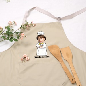 May include: Beige apron with a chef graphic and the text "Custom Text". The apron has a front pocket and a neck strap. Two wooden spatulas are next to the apron. The apron is a kitchen accessory.