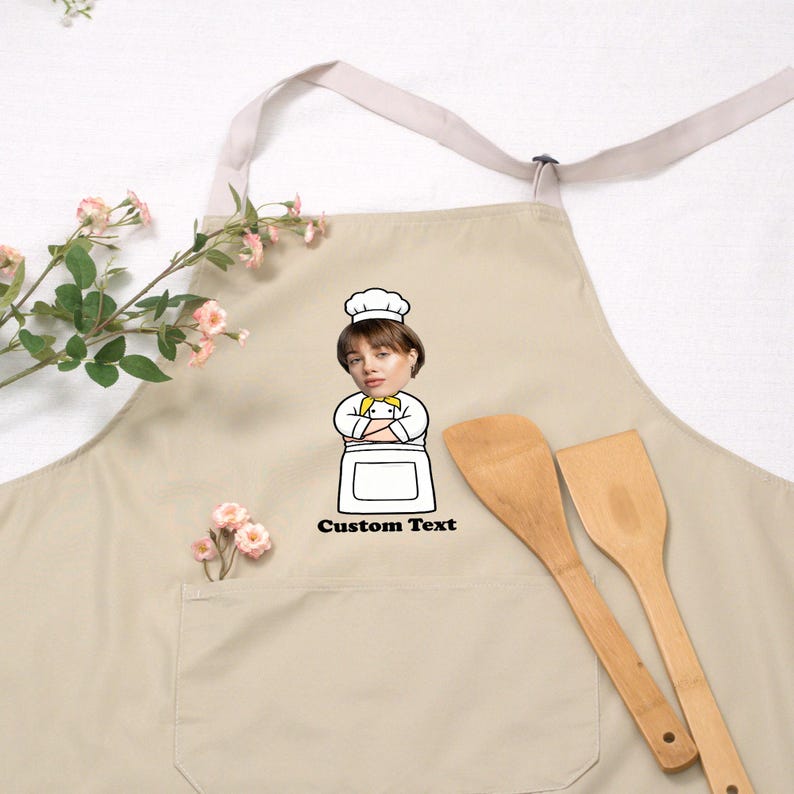 May include: Beige apron with a chef graphic and the text "Custom Text". The apron has a front pocket and a neck strap. Two wooden spatulas are next to the apron. The apron is a kitchen accessory.