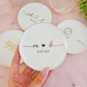 May include: White ceramic ring dishes with gold rims and personalized text. One dish has rings on it. The dishes have custom text and dates, with a heart symbol. The text includes initials and dates.