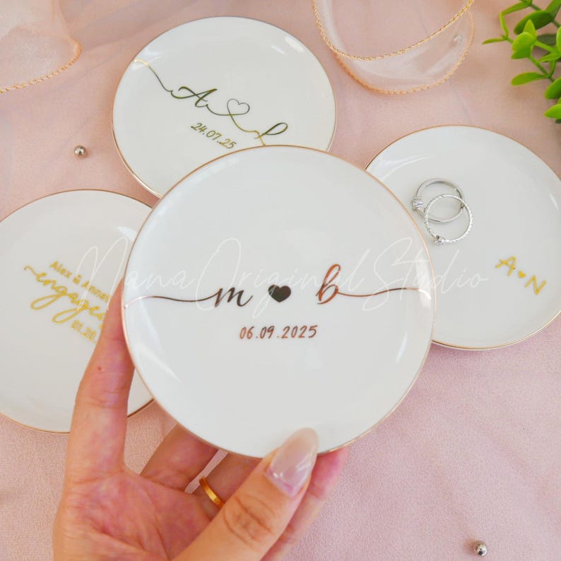 May include: White ceramic ring dishes with gold rims and personalized text. One dish has rings on it. The dishes have custom text and dates, with a heart symbol. The text includes initials and dates.