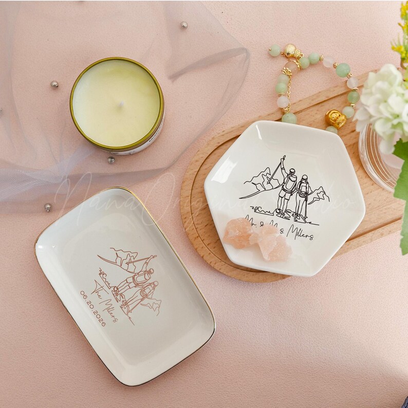 May include: Two white ceramic ring dishes with gold trim, one rectangular and one hexagonal, each featuring a mountain climbing illustration. A small candle in a gold tin and a bracelet are also visible.