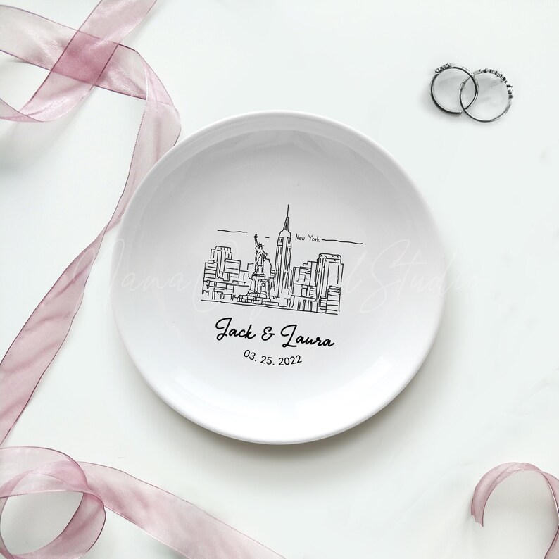 Op de afbeelding: Een witte keramische ringschaal met een zwarte lijntekening van de skyline van New York City, inclusief het Vrijheidsbeeld en het Empire State Building. De schaal is gepersonaliseerd met de namen "Jack & Laura" en de datum "03. 25. 2022". Twee zilveren ringen zijn zichtbaar.
