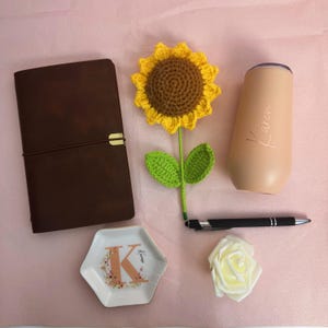 K&ouml;nnte beinhalten: Verschiedene Gegenst&auml;nde auf einer rosa Oberfl&auml;che. Ein braunes Lederjournal, eine geh&auml;kelte Sonnenblume, ein pfirsichfarbener Becher mit dem Namen "Karen", ein schwarzer Stift, eine kleine dekorative Schale mit dem Buchstaben "K" und eine wei&szlig;e Rose.