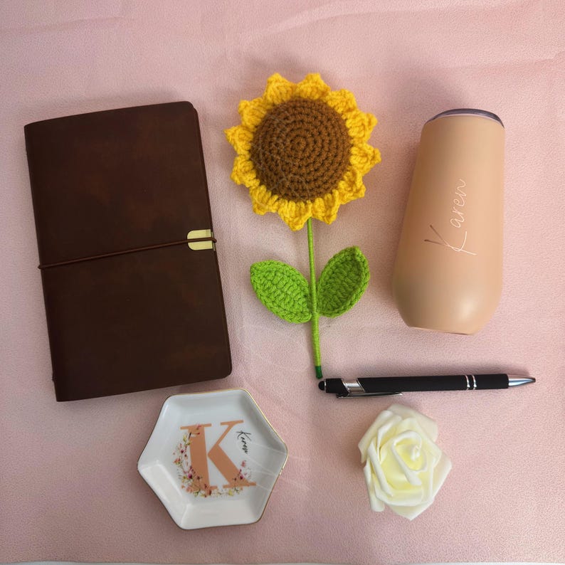 K&ouml;nnte beinhalten: Verschiedene Gegenst&auml;nde auf einer rosa Oberfl&auml;che. Ein braunes Lederjournal, eine geh&auml;kelte Sonnenblume, ein pfirsichfarbener Becher mit dem Namen "Karen", ein schwarzer Stift, eine kleine dekorative Schale mit dem Buchstaben "K" und eine wei&szlig;e Rose.