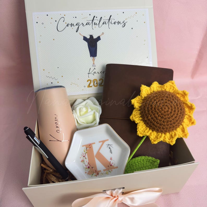 K&ouml;nnte beinhalten: Eine Geschenkbox zum Thema Abschluss, die einen Becher, einen Stift, ein Notizbuch, eine dekorative Schale und eine geh&auml;kelte Sonnenblume enth&auml;lt. Die Box enth&auml;lt auch eine Karte mit "Herzlichen Gl&uuml;ckwunsch" und eine wei&szlig;e Rose. Der Becher und die Schale sind mit einem Namen personalisiert.