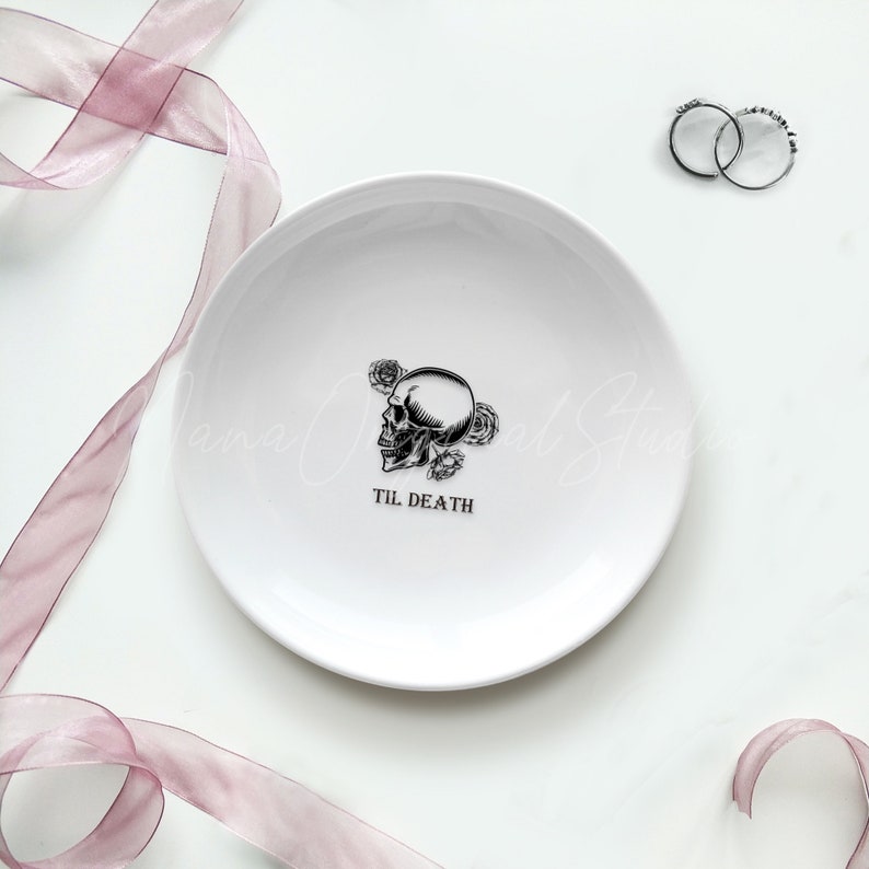 May include: A round white ceramic trinket dish featuring a black graphic of a skull surrounded by roses. The text TIL DEATH is printed in black below the skull. Two silver rings are positioned near the top right edge of the dish.