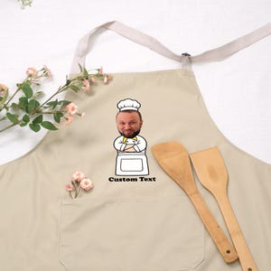 May include: Beige apron with a chef graphic and the text "Custom Text". The apron has a pocket and a light-colored neck strap. Two wooden spatulas are next to the apron.