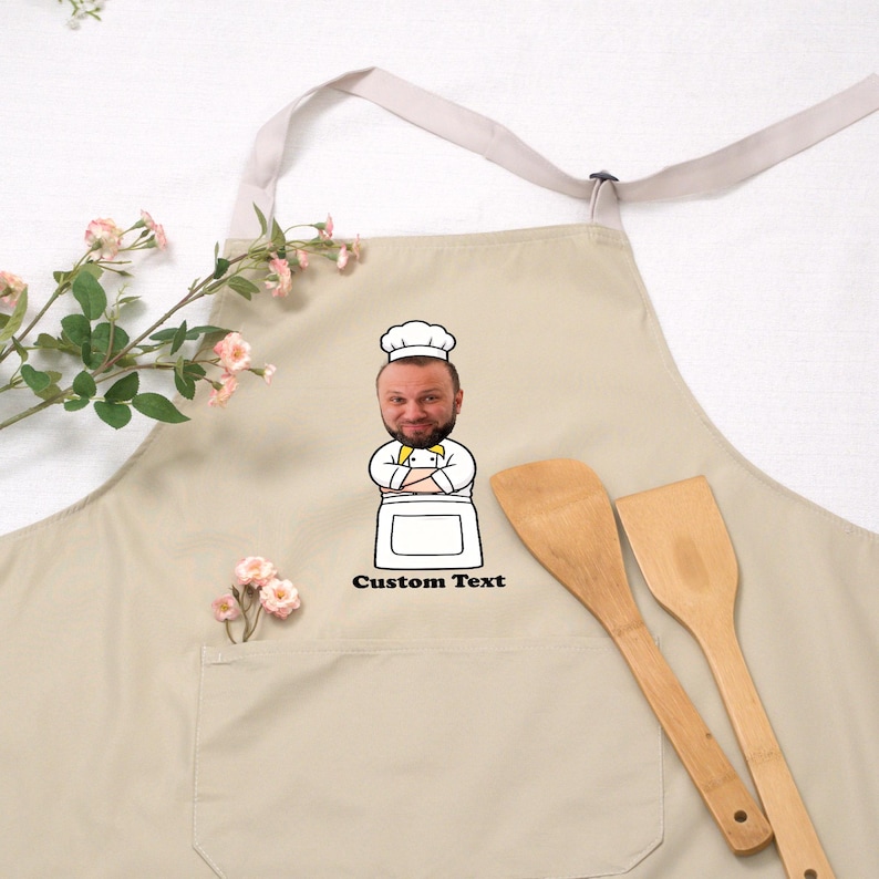 May include: Beige apron with a chef graphic and the text "Custom Text". The apron has a pocket and a light-colored neck strap. Two wooden spatulas are next to the apron.