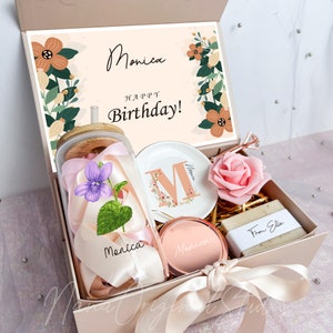 May include: A gift box with a personalized theme. Inside, a glass jar with a straw, a decorative plate with the letter "M", a compact mirror, a rose, and a bar of soap. The box includes a card that says "Monica Happy Birthday!"