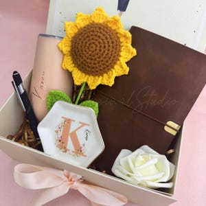 K&ouml;nnte beinhalten: Geschenkbox mit einem braunen Lederjournal, einem beigen Becher mit dem Namen "Karon", einem schwarzen Stift, einer wei&szlig;en Blumenschale mit dem Buchstaben "K", einer geh&auml;kelten Sonnenblume und einer wei&szlig;en Rose. Die Box ist mit einem rosa Band gebunden.