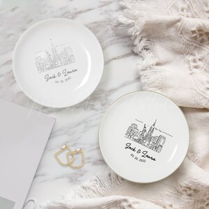 Op de afbeelding: Twee witte keramische ring schaaltjes met een zwarte lijntekening van de skyline van New York City. Elk schaaltje is gepersonaliseerd met de namen "Jack & Laura" en de datum "03. 25. 2022". E&eacute;n schaaltje heeft een gouden rand.