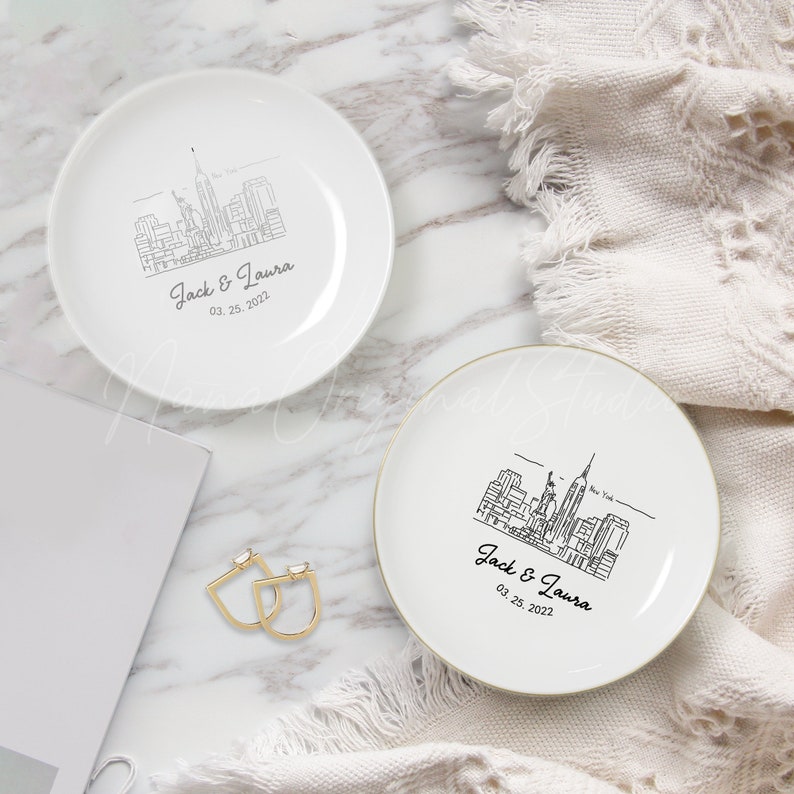 Op de afbeelding: Twee witte keramische ring schaaltjes met een zwarte lijntekening van de skyline van New York City. Elk schaaltje is gepersonaliseerd met de namen "Jack & Laura" en de datum "03. 25. 2022". E&eacute;n schaaltje heeft een gouden rand.
