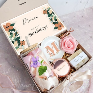 May include: A gift box with a variety of items, including a glass tumbler with a flower and ribbon, a small dish with the letter "M", a rose, a compact mirror, and a bar of soap. The box lid has the words "Monica Happy Birthday!"