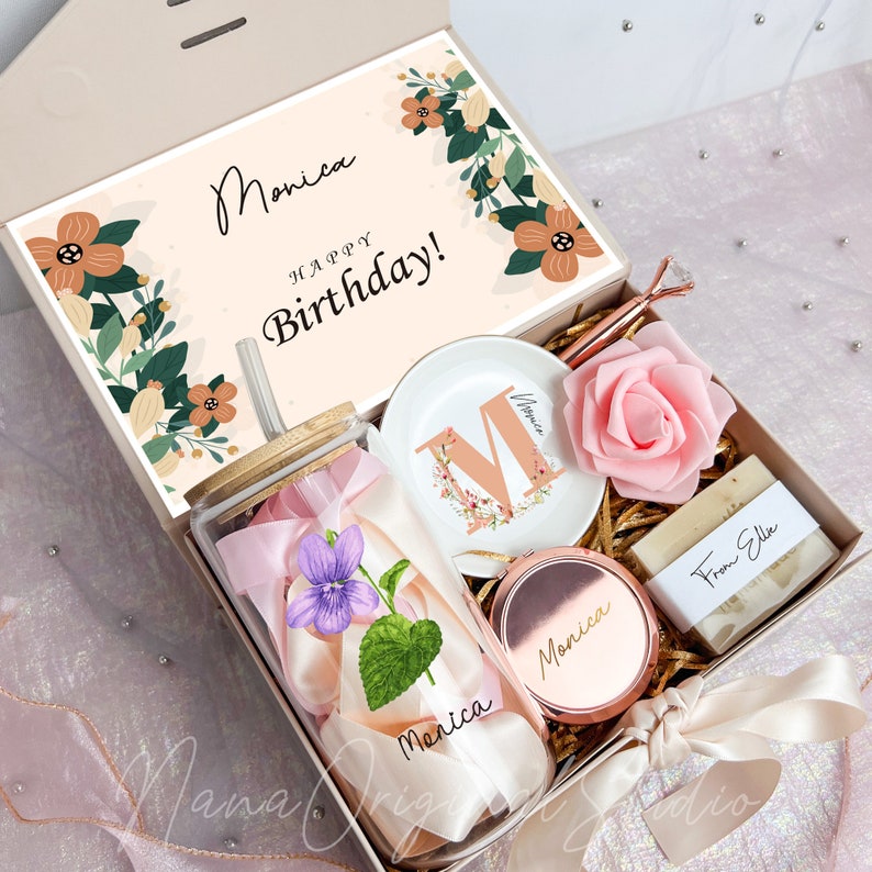 May include: A gift box with a variety of items, including a glass tumbler with a flower and ribbon, a small dish with the letter "M", a rose, a compact mirror, and a bar of soap. The box lid has the words "Monica Happy Birthday!"
