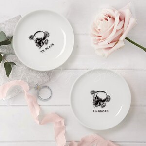 May include: Two white ceramic ring dishes, each featuring a black graphic of a human skull surrounded by roses and the text TIL DEATH printed below. The dishes are round with a smooth finish and are presented alongside a silver ring.