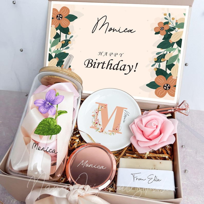 May include: A gift box with a variety of items, including a glass jar with a flower and ribbon, a decorative plate with the letter "M", a compact mirror, a rose, and a bar of soap. The box has the name "Monica" and the words "Happy Birthday!"