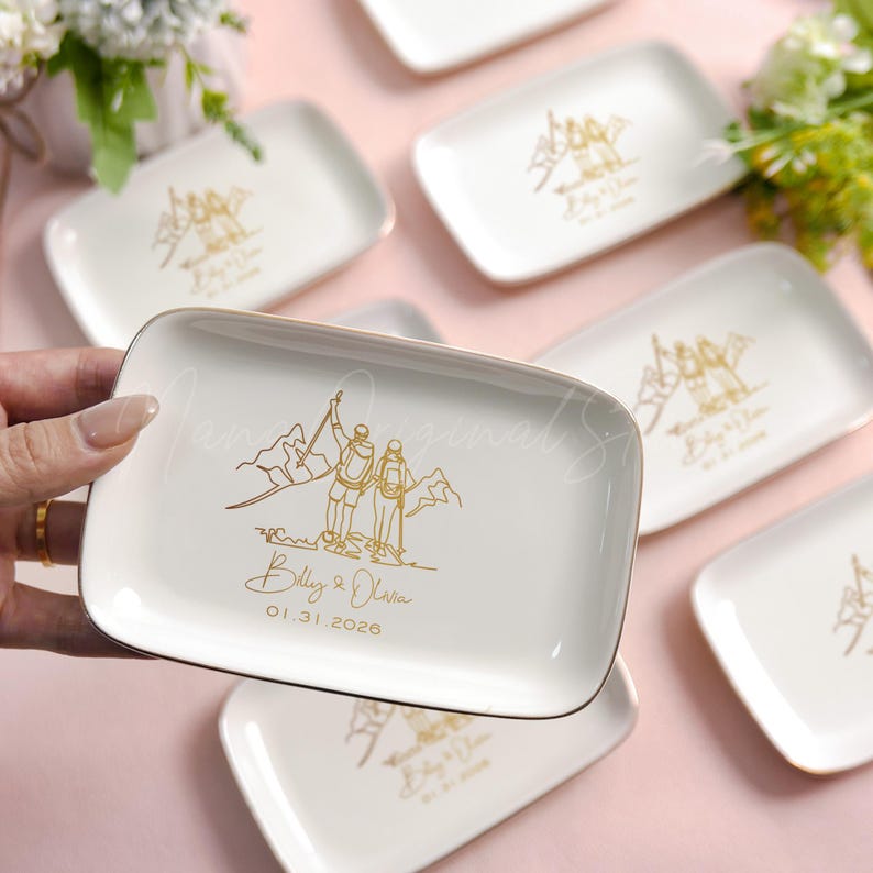 May include: Rectangular white ceramic trinket trays with gold trim. Each tray features a gold illustration of two people on a mountain, with the names "Billy & Olivia" and the date "01.31.2026" below. The trays are arranged on a pink surface.