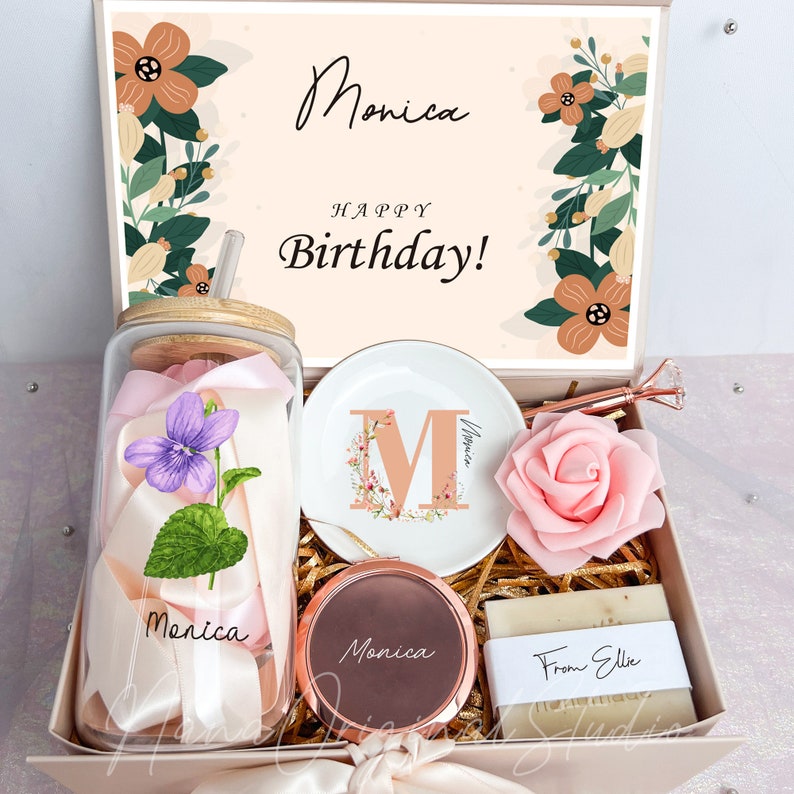 May include: A gift box with a personalized theme. It contains a glass jar with a straw, a decorative plate with the letter "M", a compact mirror, a rose, and soap. The box is decorated with floral elements and the text "Monica Happy Birthday!"