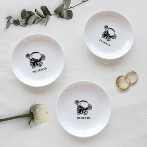 May include: Three white ceramic trinket dishes, each featuring a black graphic of a skull surrounded by roses and the text "TIL DEATH" printed in the center. The dishes are arranged with two gold rings and a white flower nearby.