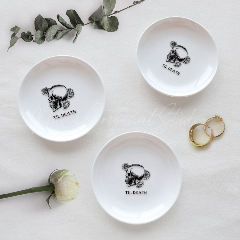 May include: Three white ceramic trinket dishes, each featuring a black graphic of a skull surrounded by roses and the text "TIL DEATH" printed in the center. The dishes are arranged with two gold rings and a white flower nearby.