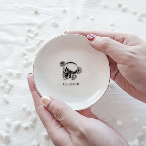 May include: A round white ceramic trinket dish with a gold-toned rim. The center features a black graphic illustration of a human skull surrounded by small roses, with the text TIL DEATH printed in a bold black serif font below the skull.