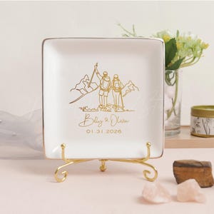 May include: A square, white ceramic trinket dish with a gold rim and a gold stand. The dish features a gold line drawing of a couple hiking in the mountains, with the names "Billy & Olivia" and the date "01.31.2026" below.