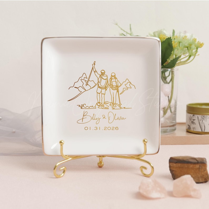 May include: A square, white ceramic trinket dish with a gold rim and a gold stand. The dish features a gold line drawing of a couple hiking in the mountains, with the names "Billy & Olivia" and the date "01.31.2026" below.