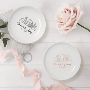 Op de afbeelding: Twee witte keramische borden met een ontwerp van de skyline van Los Angeles. E&eacute;n bord heeft een zwart ontwerp, de andere een koperkleurig ontwerp. Beide borden zijn voorzien van de namen "Danielle & Ashley" en de datum "02.27.2022". Een roze roos en een ring zijn ook zichtbaar.