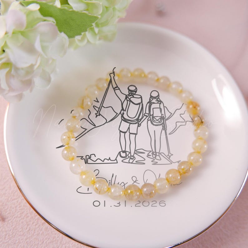 May include: A beaded bracelet with yellow and clear stones rests on a white plate with a gold rim. The plate features a black illustration of two hikers and the date 01.31.2026. White flowers are in the background.