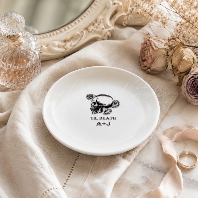 May include: A white ceramic trinket dish featuring a black graphic of a skull surrounded by roses. The text "TIL DEATH" is printed above the initials "A+J" in a black serif font centered on the surface of the round dish.