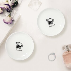 May include: Two round white ceramic trinket dishes, each featuring a black graphic of a human skull surrounded by roses and the text TIL DEATH printed below. The dishes are smooth with a glossy finish and a simple, clean design.