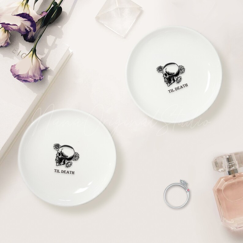 May include: Two round white ceramic trinket dishes, each featuring a black graphic of a human skull surrounded by roses and the text TIL DEATH printed below. The dishes are smooth with a glossy finish and a simple, clean design.