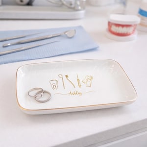 Op de afbeelding: Een wit rechthoekig keramisch dienblad met gouden rand, met gouden illustraties van tandheelkundige instrumenten en de naam "Ashley". Twee zilveren ringen liggen op het dienblad. Tandheelkundige instrumenten en een tandmodel op de achtergrond.