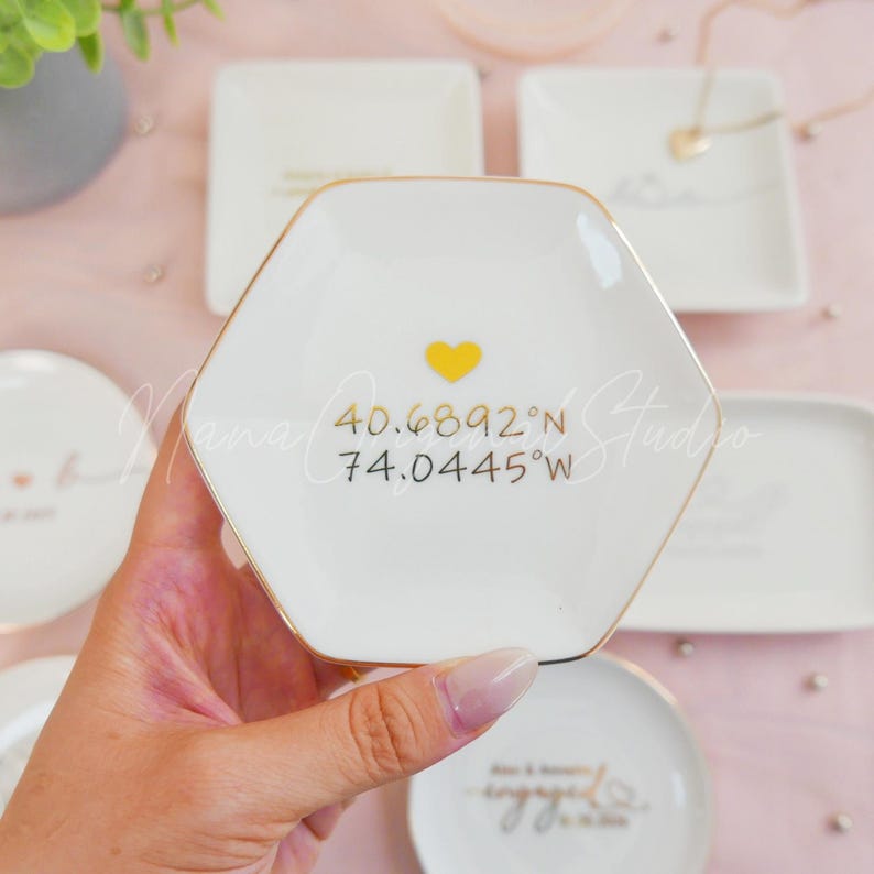 May include: White ceramic hexagonal trinket dish with a gold rim, a yellow heart, and coordinates 40.6892&deg;N 74.0445&deg;W. Other white ceramic dishes are in the background.