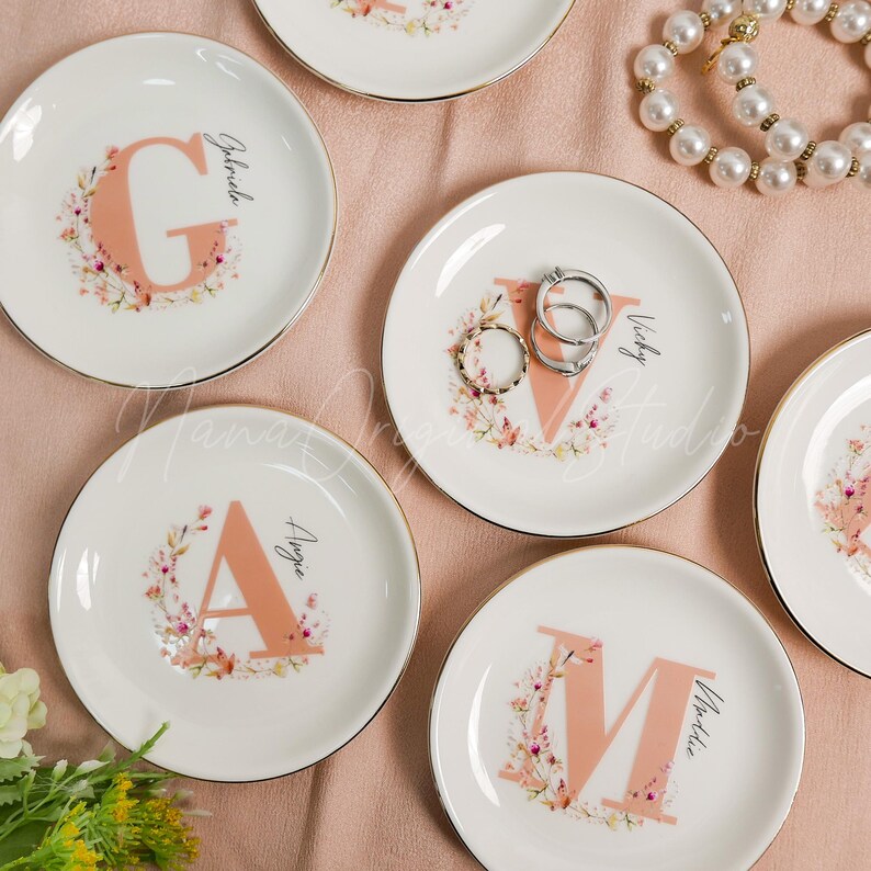 May include: A collection of round white ceramic trinket dishes with gold-toned rims. Each dish features a large peach-colored letter surrounded by a delicate floral wreath and a name in black script. Two silver rings rest on the dish with the letter V.