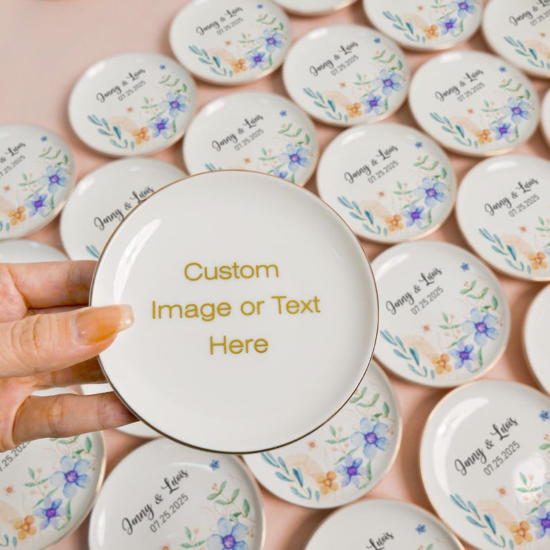 May include: A round white ceramic trinket dish with a gold rim. The center features the gold text 'Custom Image or Text Here'. Other identical dishes in the background display a floral watercolor design with the text 'Jinny & Luis 07.25.2025'.
