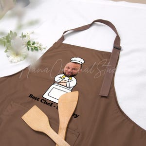 May include: A brown apron with a cartoon chef graphic and the text "Best Chef - [Name]" is displayed. Two wooden spatulas are in front of the apron. The apron has a pocket and a brown neck strap.