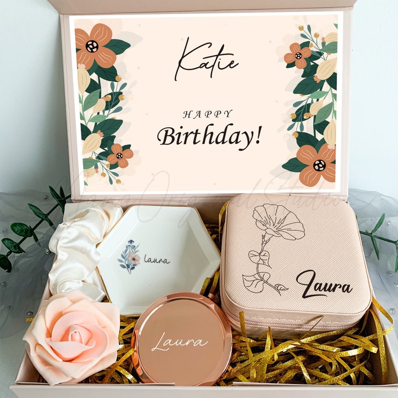 Puede incluir: Una caja de regalo rosa con un dise&ntilde;o floral y el texto "Katie &iexcl;Feliz cumplea&ntilde;os!" en la tapa. La caja contiene un plato de anillo blanco con el nombre "Laura" en &eacute;l, una caja de joyer&iacute;a rosa con el nombre "Laura" en ella, un espejo compacto de oro rosa con el nombre "Laura" en &eacute;l y una scrunchie de sat&eacute;n blanco.