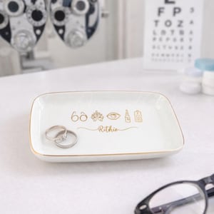 Könnte beinhalten: Ein weißes, rechteckiges Keramiktablett mit goldenem Rand, das goldfarbene Symbole für die Augenpflege und den Namen "Rithie" zeigt. Zwei silberne Ringe liegen auf dem Tablett. Im Hintergrund befinden sich eine Sehtafel und Augenuntersuchungsgeräte.