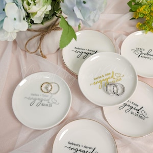 May include: White ceramic ring dishes with gold rims and the word "engaged" in various fonts. Each dish has a couple's names and engagement date. Some dishes hold rings. Floral arrangement in the background.