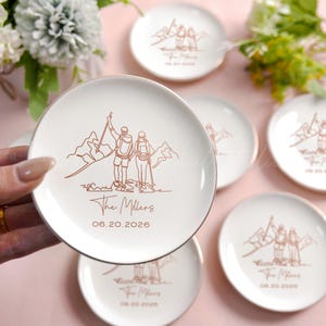 May include: White ceramic plates with a gold rim feature a line drawing of a couple hiking in the mountains. The text "The Millers" and the date "06.20.2026" are below the illustration. Several plates are visible.