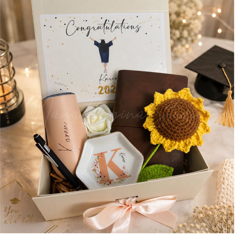 K&ouml;nnte beinhalten: Eine Geschenkbox zur Graduierung mit einem personalisierten Becher, einem Stift, einem Notizbuch und einer geh&auml;kelten Sonnenblume. Die Box enth&auml;lt eine dekorative Schale mit dem Buchstaben "K" und eine Karte mit der Aufschrift "Herzlichen Gl&uuml;ckwunsch". Die Box ist mit einer Pfirsichschleife gebunden.