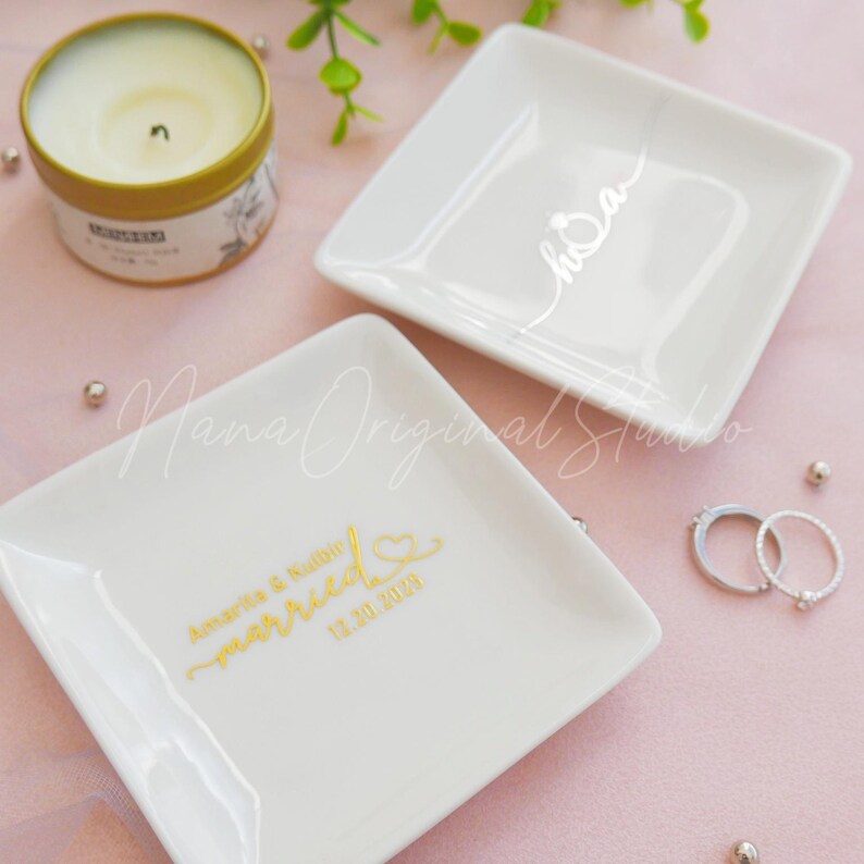 May include: Two white square ring dishes with gold lettering. One dish says "married" with a date and names, the other says "hoa". A gold candle and rings are also in the image. The dishes are on a pink surface.