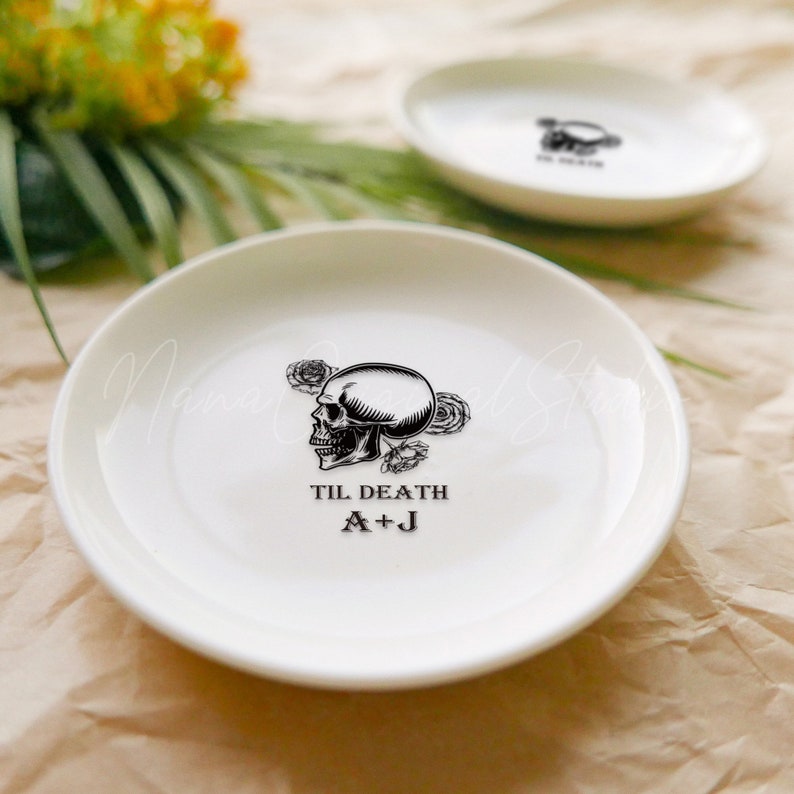 May include: Two white ceramic ring dishes featuring a black graphic of a skull surrounded by roses. The text TIL DEATH and the initials A+J are printed in black below the skull illustration on the front-most dish.