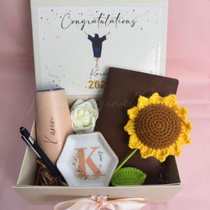 K&ouml;nnte beinhalten: Eine Geschenkbox zum Thema Abschluss, die einen Becher, einen Stift, ein Notizbuch, eine dekorative Schale und eine geh&auml;kelte Sonnenblume enth&auml;lt. Die Box enth&auml;lt auch eine Karte mit der Aufschrift "Herzlichen Gl&uuml;ckwunsch" sowie den Namen "Karen" und das Jahr "2023".