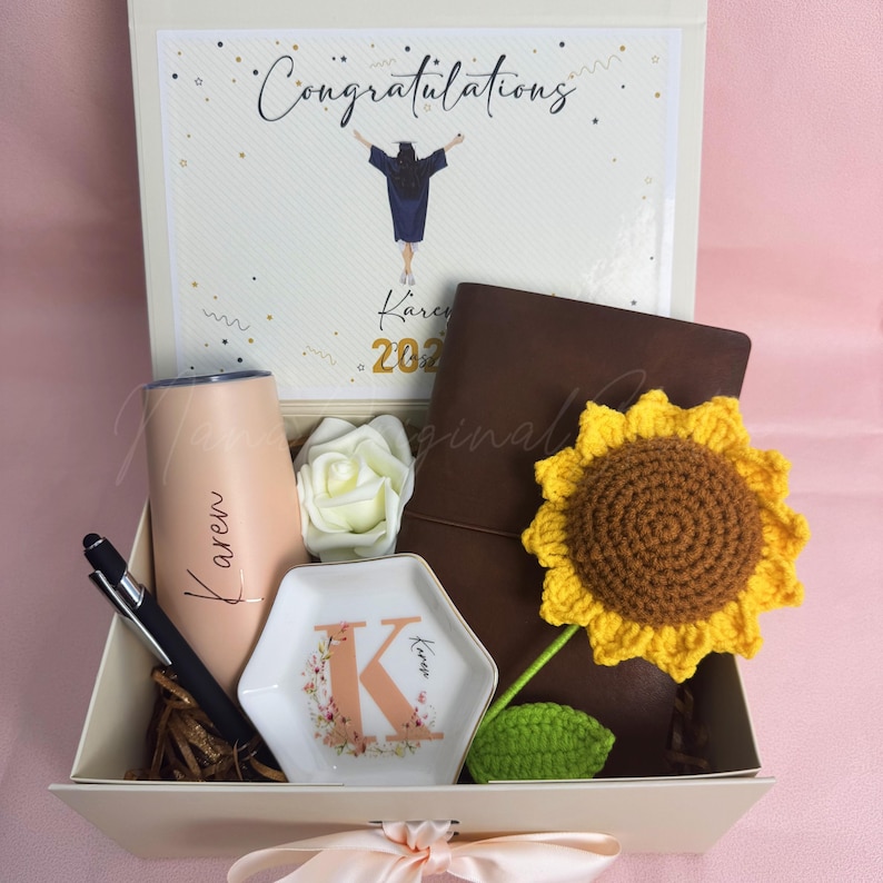 K&ouml;nnte beinhalten: Eine Geschenkbox zum Thema Abschluss, die einen Becher, einen Stift, ein Notizbuch, eine dekorative Schale und eine geh&auml;kelte Sonnenblume enth&auml;lt. Die Box enth&auml;lt auch eine Karte mit der Aufschrift "Herzlichen Gl&uuml;ckwunsch" sowie den Namen "Karen" und das Jahr "2023".