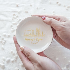 Op de afbeelding: Een witte keramische ringenschaal met een gouden rand, met een gouden skyline-ontwerp en de namen "Harvey & Sophia" met de datum "06.20.2022". De schaal wordt in twee handen gehouden.